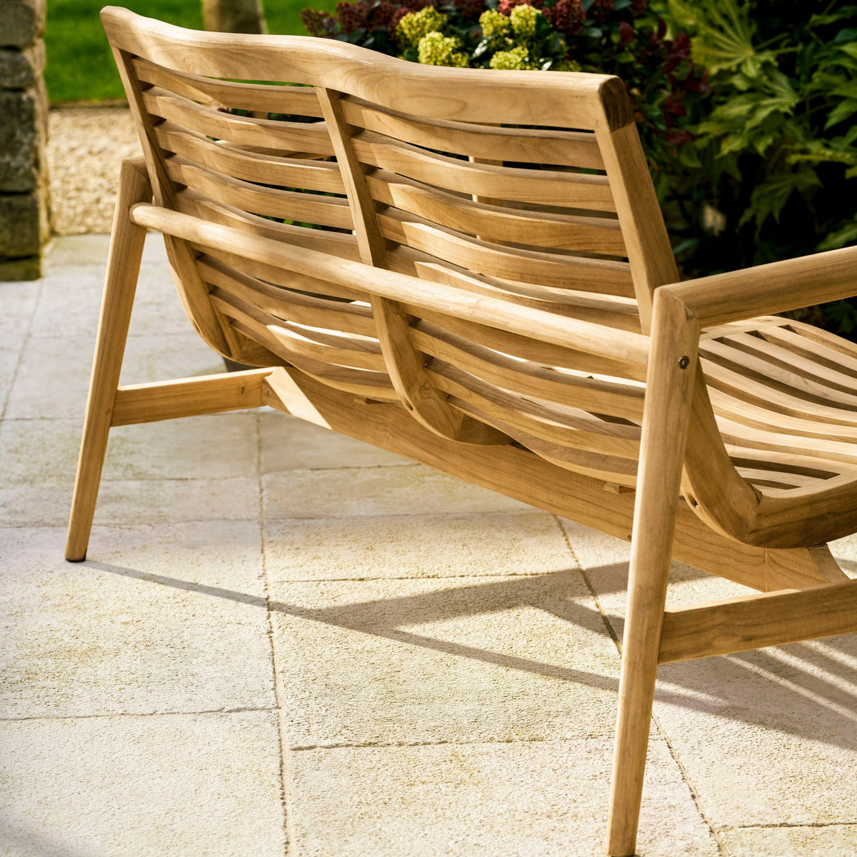 A light natural wood outdoor lounge chair with curved slatted back and seat, shown on a stone patio in a garden setting.