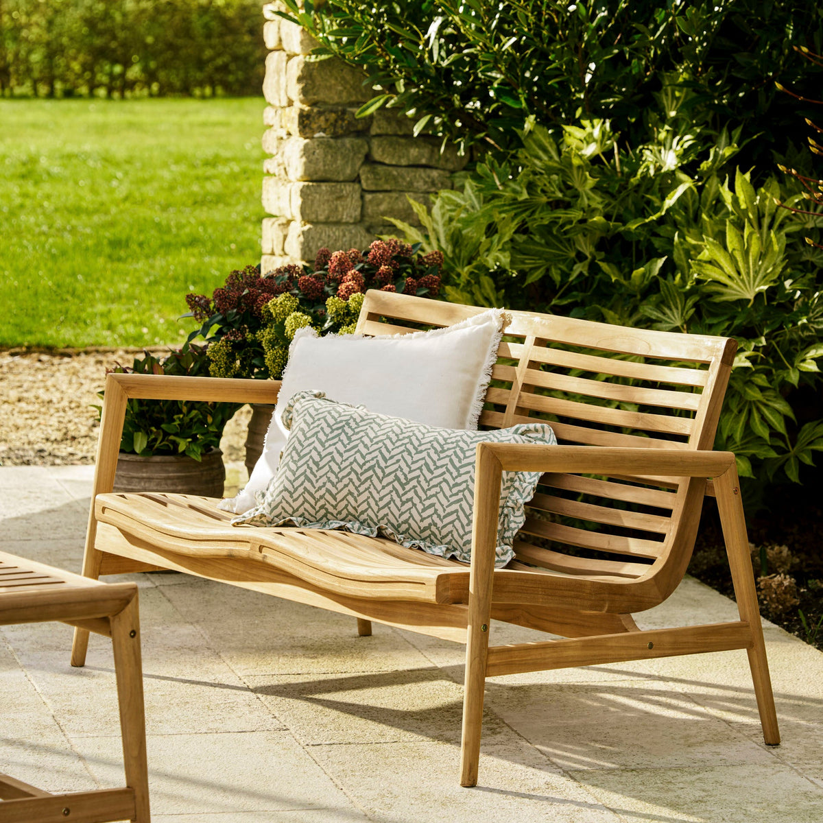 A wooden outdoor patio loveseat with two decorative throw pillows, set on a stone patio in a landscaped garden with green grass and shrubs.