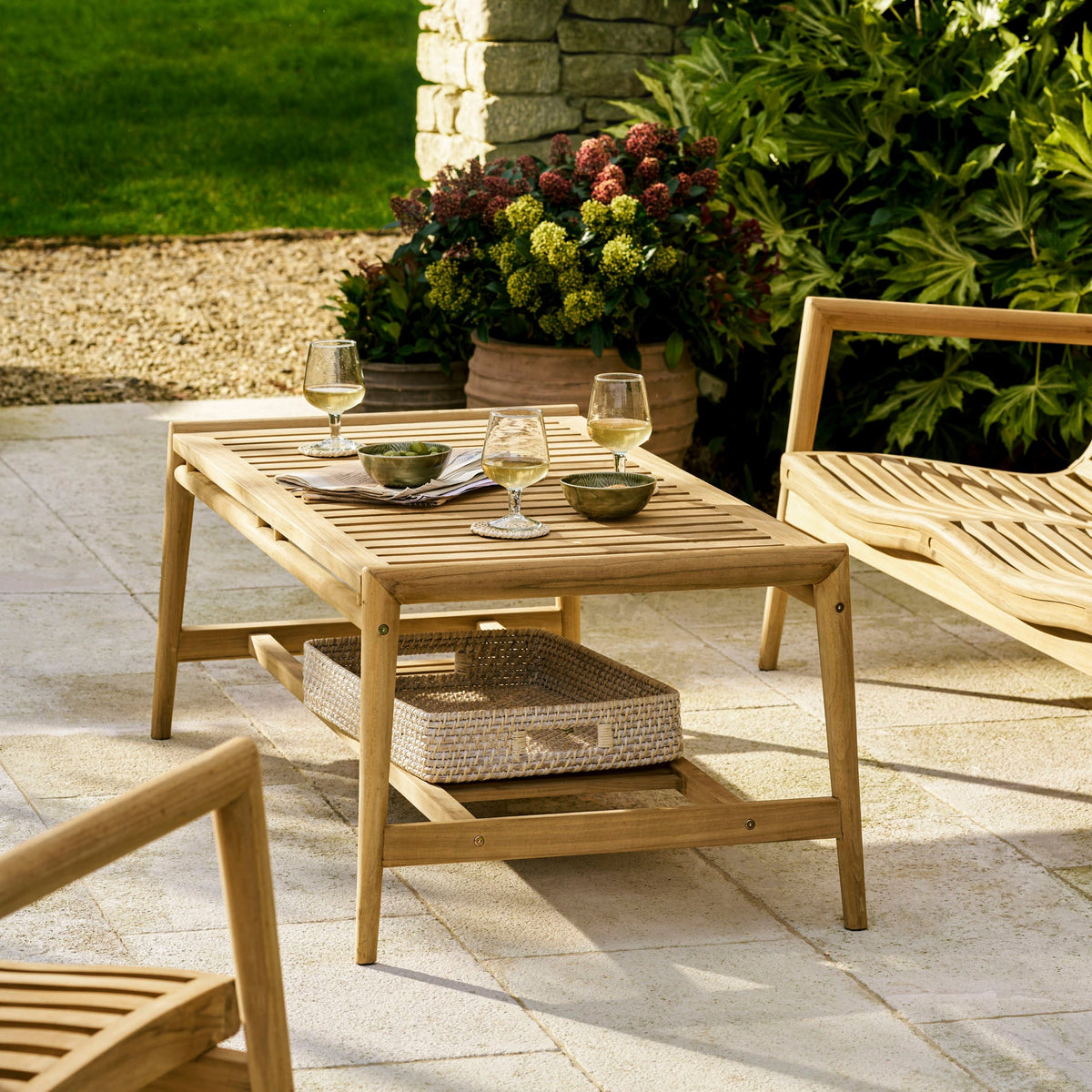 A light natural wood outdoor patio coffee table with a slatted top and lower shelf, styled on a stone patio with chairs, wine glasses, and potted plants in a garden setting.