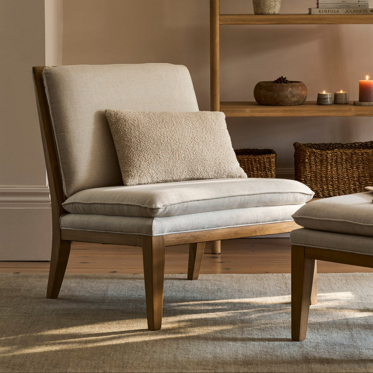A light beige upholstered accent chair with a wooden frame and a matching textured throw pillow, styled in a warm, neutral-toned room with a wooden bookshelf in the background.