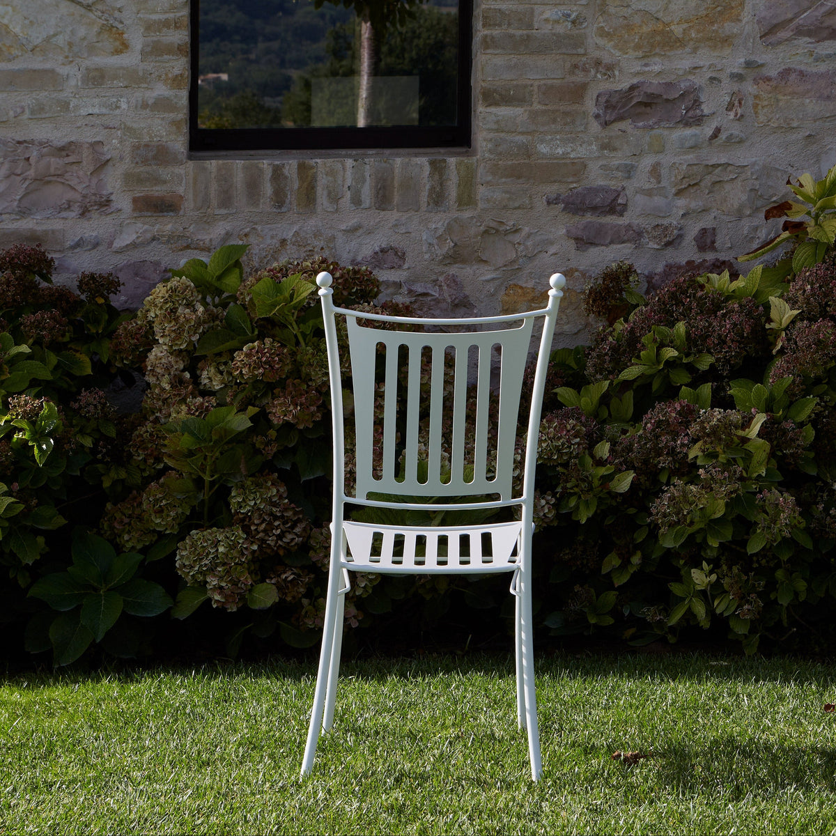 A white metal garden chair with a slatted back and seat, positioned on a green lawn in front of a stone wall and leafy green shrubs.