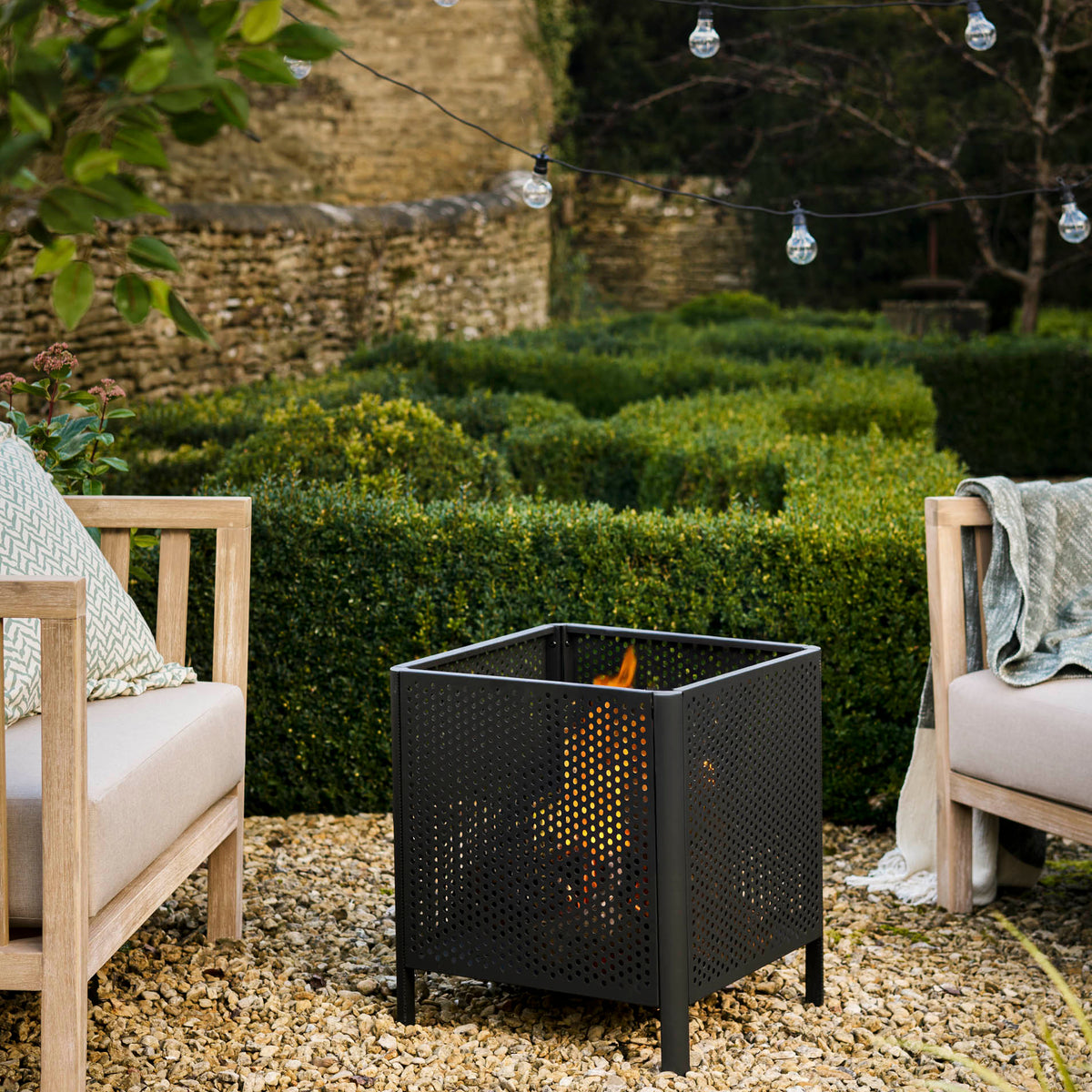 A black metal square fire pit with a perforated mesh design, containing a fire, placed on a gravel patio between two outdoor wooden armchairs in a garden setting.