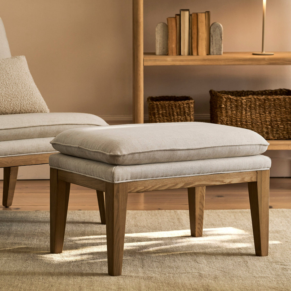 A light beige upholstered ottoman with a wooden frame and tapered legs, shown in a styled living room setting next to a matching armchair and in front of a wooden bookshelf.