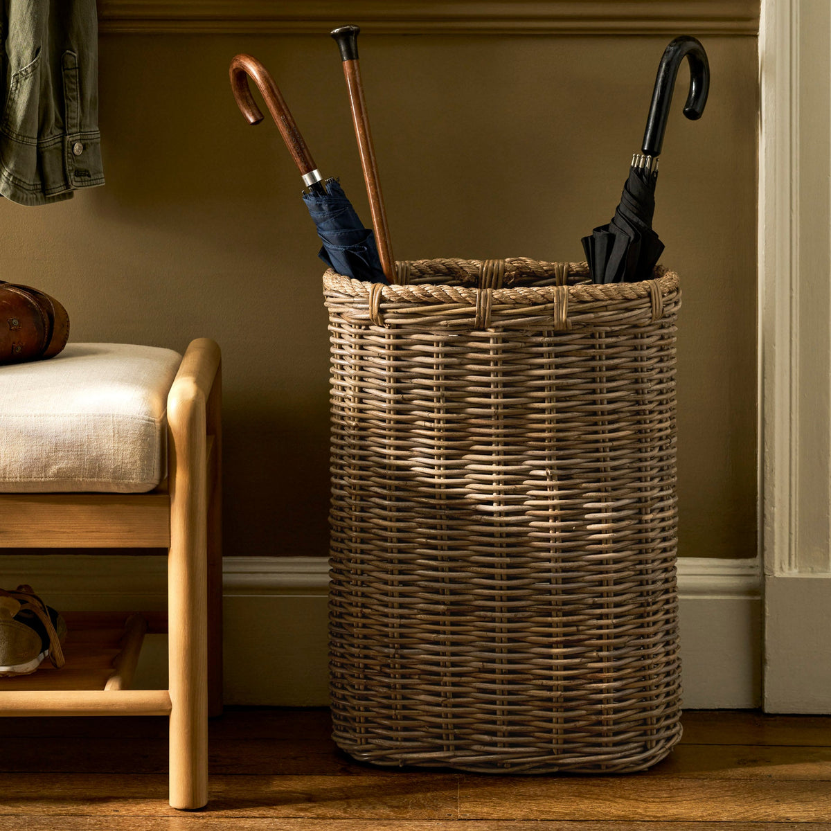 A tall woven wicker basket stand holding three umbrellas and one cane, placed on a wooden floor against a brown wall with molding and a bench visible.