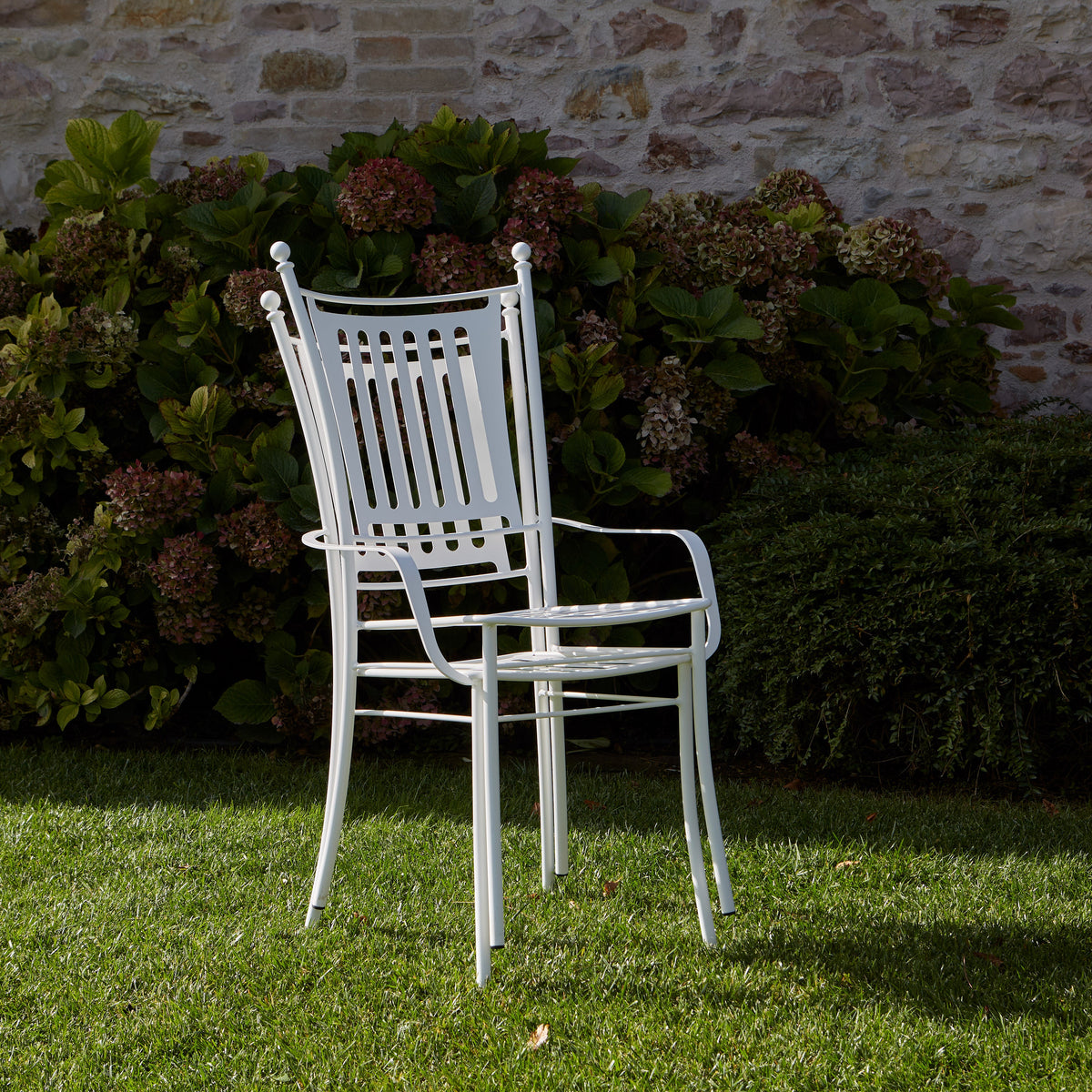 Two white metal garden chair swith a slatted backs and armrests, stacked on top of eachother and standing on a green lawn in front of a stone wall and leafy shrubs.