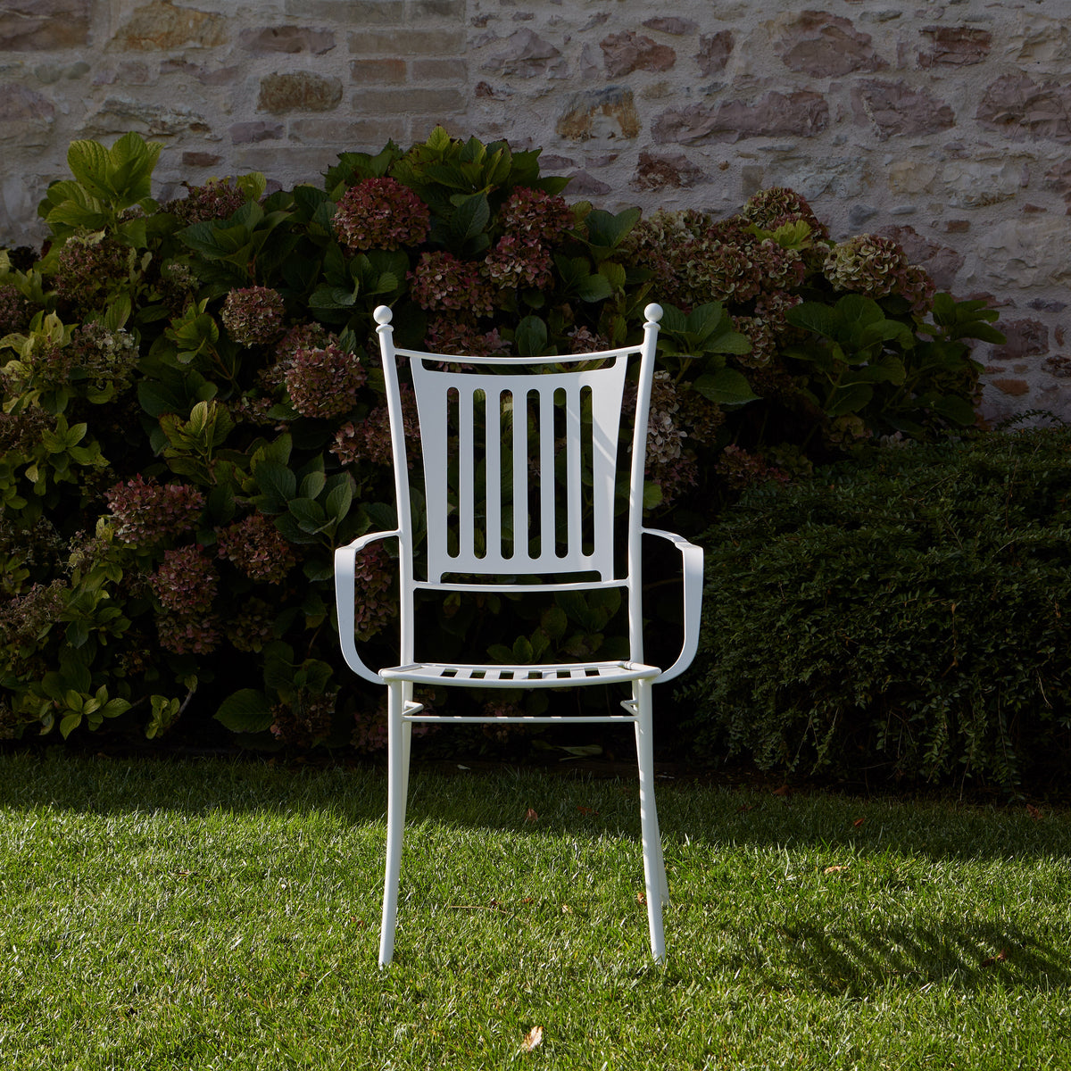 A white metal garden chair with armrests is positioned on a green lawn in front of a stone wall and leafy green shrubs.