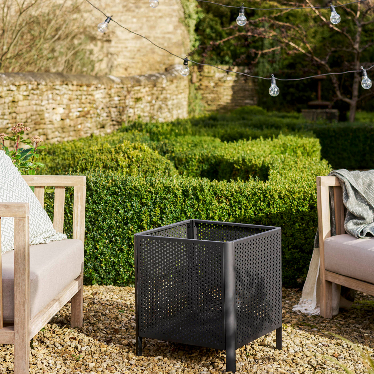 A black metal outdoor fire pit with a perforated wire design sits on a gravel patio between two wooden lounge chairs in a garden setting with hedges and string lights.
