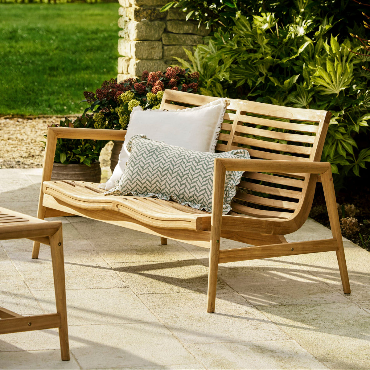 A wooden outdoor patio loveseat with two decorative throw pillows, set on a light stone patio in a landscaped garden with green grass and shrubs.