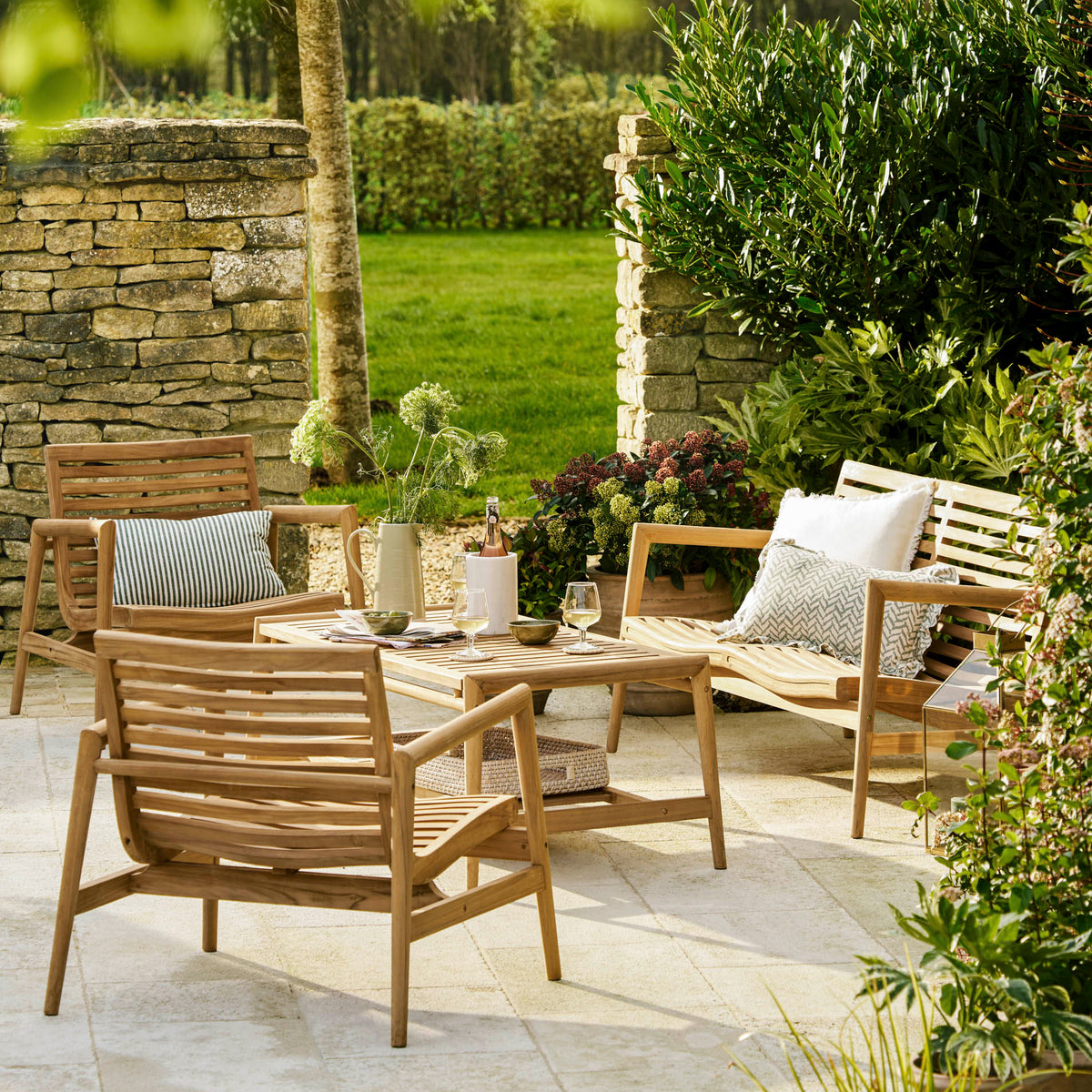 Outdoor patio furniture set in a landscaped garden, featuring a wooden slat lounge chair, armchair, bench, and matching coffee table on a light stone paver surface.