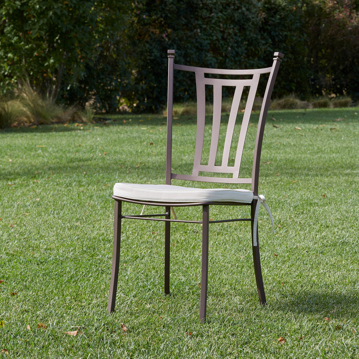 A single outdoor chair with a light beige seat cushion and a dark brown metal frame, set on a green lawn with trees in the background.