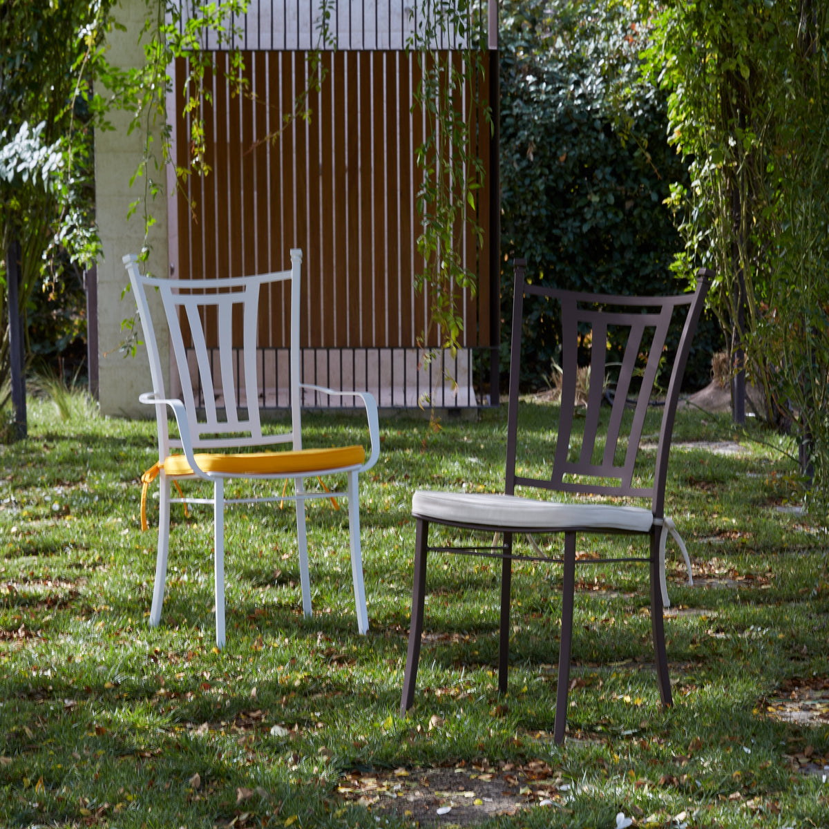 Two outdoor patio chairs on a grassy lawn surrounded by trees. The left chair is white with a yellow seat cushion, and the right chair is dark gray with a light beige seat cushion.