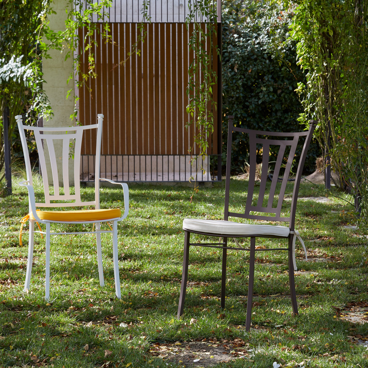 Two outdoor dining chairs on a grassy lawn surrounded by trees. The left chair is white with a yellow seat cushion, and the right chair is dark gray with a light beige seat cushion.