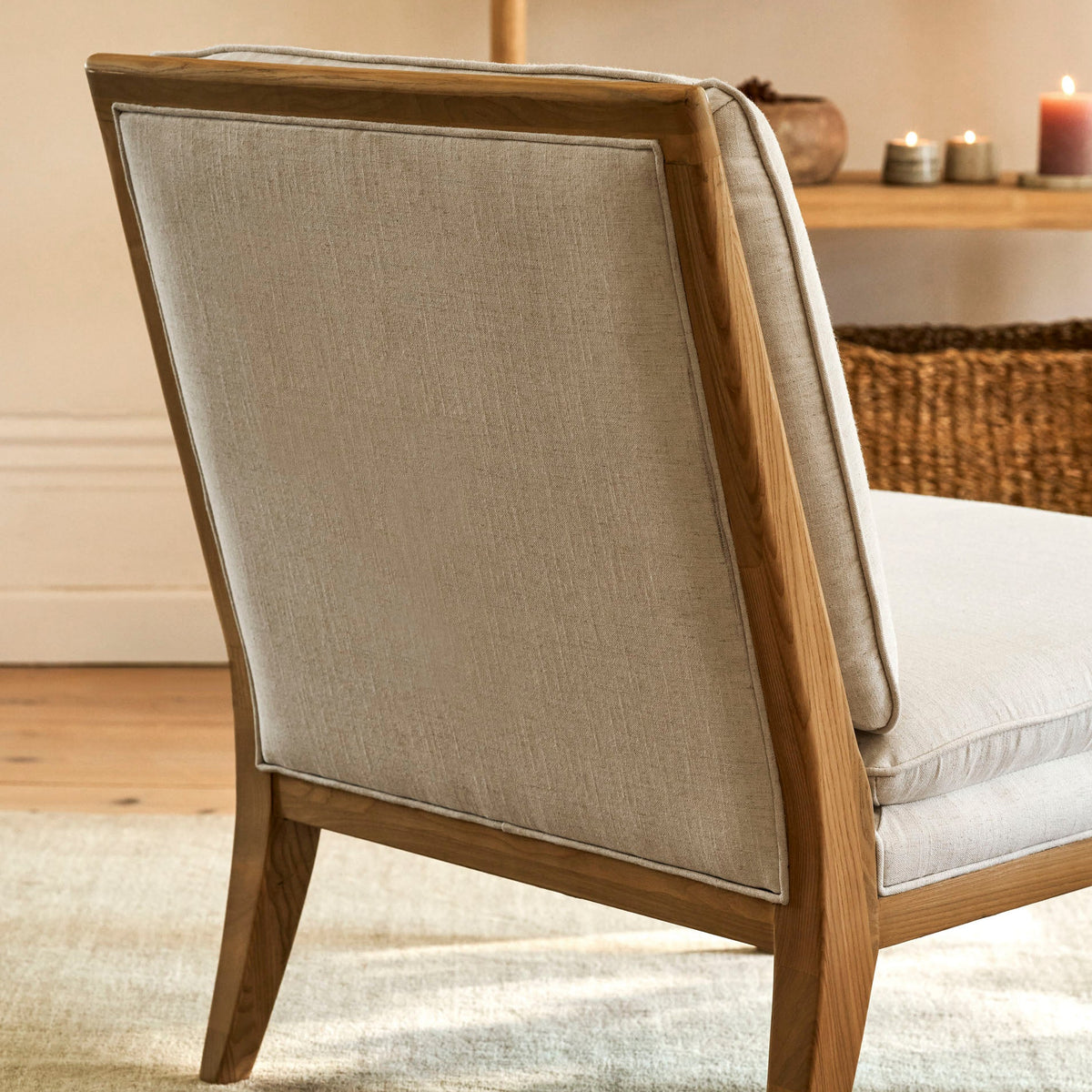 A close-up of a modern accent chair featuring a light beige linen fabric upholstery and a medium brown wooden frame, set on a textured beige rug in a warm, softly lit room.