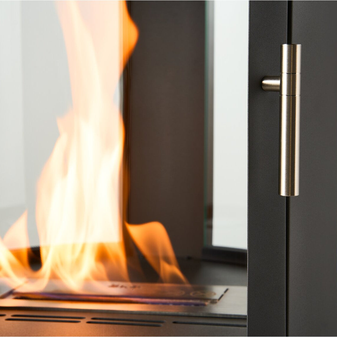 Close-up of a modern black fireplace or stove with visible orange and yellow flames rising from the firebox, alongside a brushed metal handle on the right side.