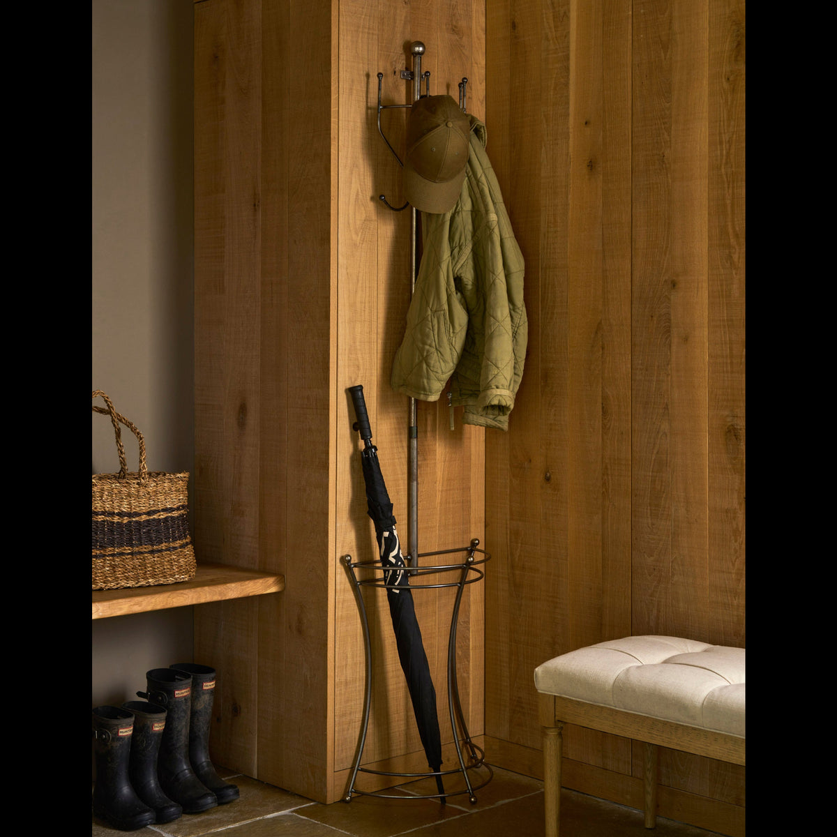 A tall wooden entryway corner with a black metal umbrella stand holding a closed black umbrella, a hat hanging on wall hooks, and an olive quilted jacket draped over the stand. A wooden bench with a cream cushion sits on the right, and boots and a woven basket are visible on the left.