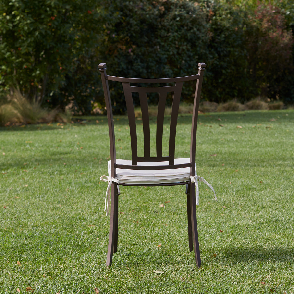 A dark brown stainless steel chair with a white cushioned seat and tied white ribbons, photographed outdoors on a green lawn with trees in the background
