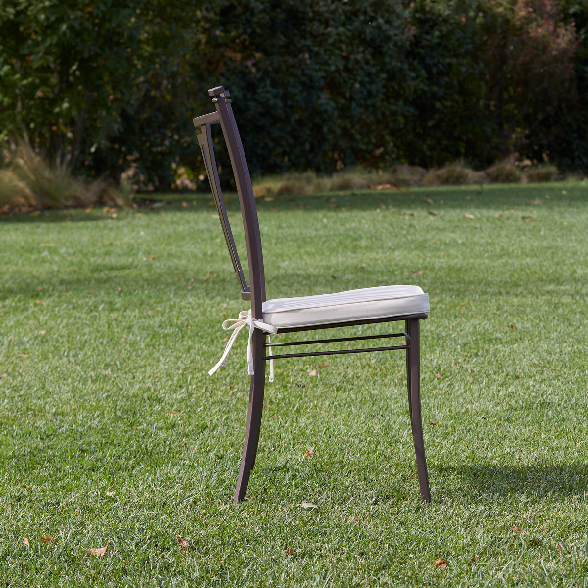A dark brown stainless steel dining chair with a light beige cushion and white bow tie sits on a green grass lawn with a blurred garden background.