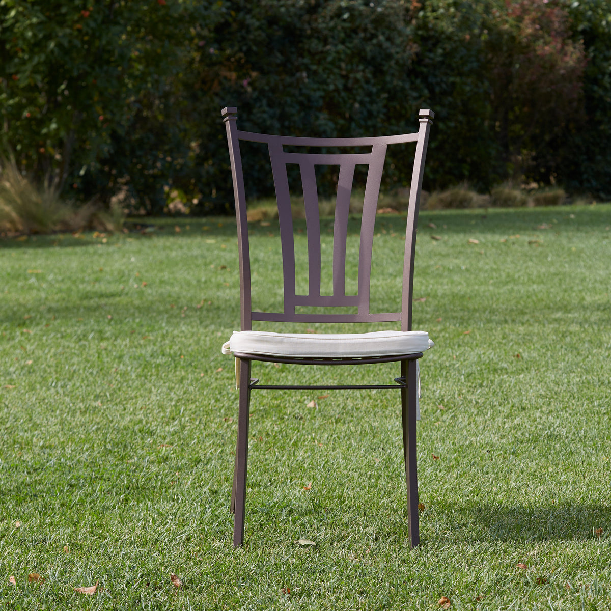 A single stainless steel dining chair with a light beige upholstered seat and a dark brown frame, positioned on a green grass lawn with a background of trees and shrubs.