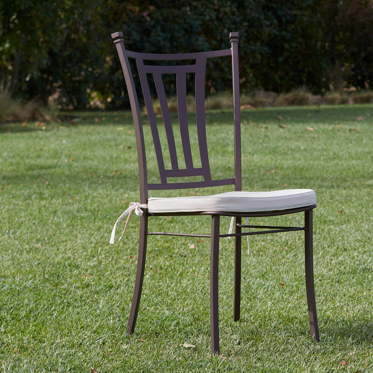 A dark metal outdoor dining chair with a white seat cushion tied with a ribbon, set on a green lawn with trees in the background.