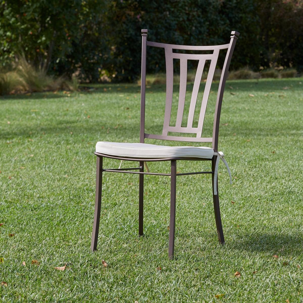 A single outdoor chair with a light beige seat cushion and a dark brown metal frame, set on a green lawn with trees in the background.