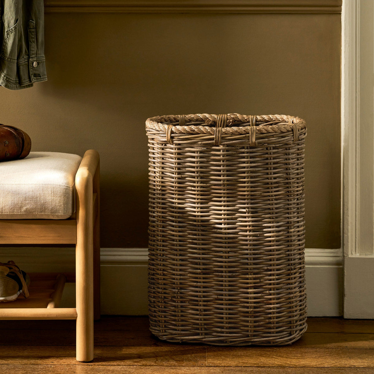 A tall, cylindrical woven wicker laundry hamper in a natural tan color, positioned on a wooden floor next to a light wood bench with a white cushion in a warm, rustic room setting.