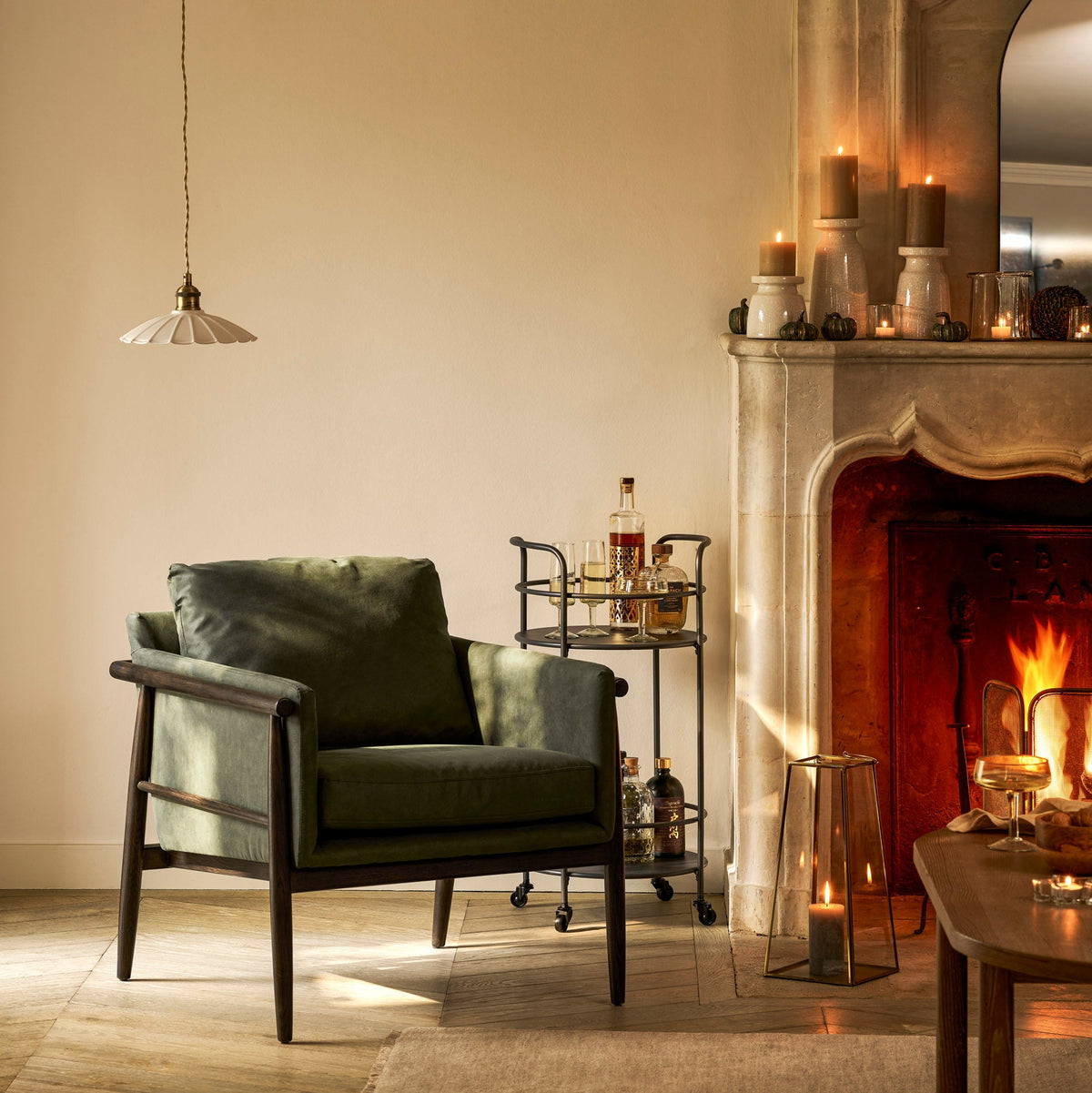 A dark green upholstered armchair with dark wood arms and legs, styled in a cozy room next to a stone fireplace with candles and a warm fire.