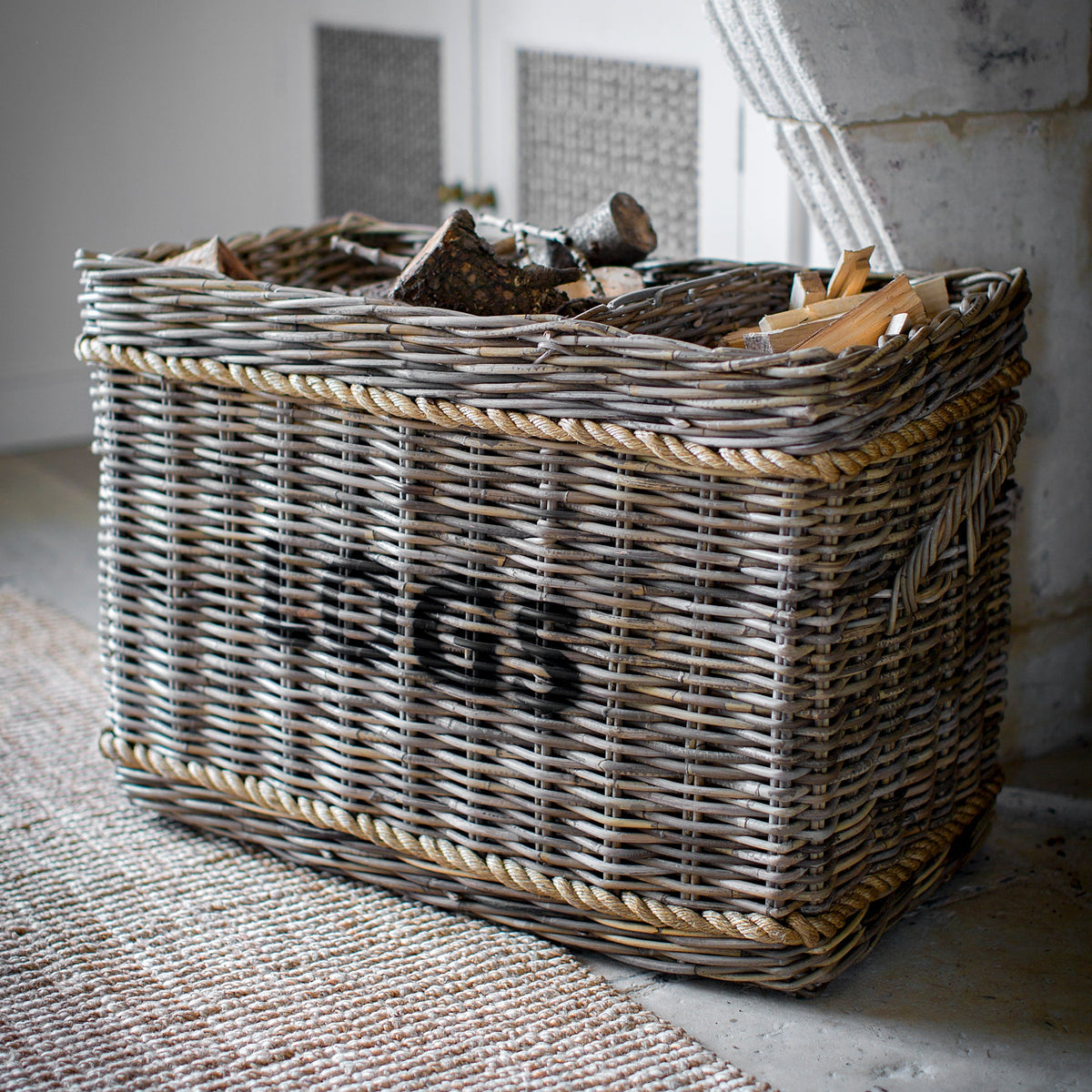 A rectangular woven wicker storage basket filled with firewood, placed on a textured rug in a rustic indoor setting. The basket has a black stencil-style logo reading 'JGS' on the front.