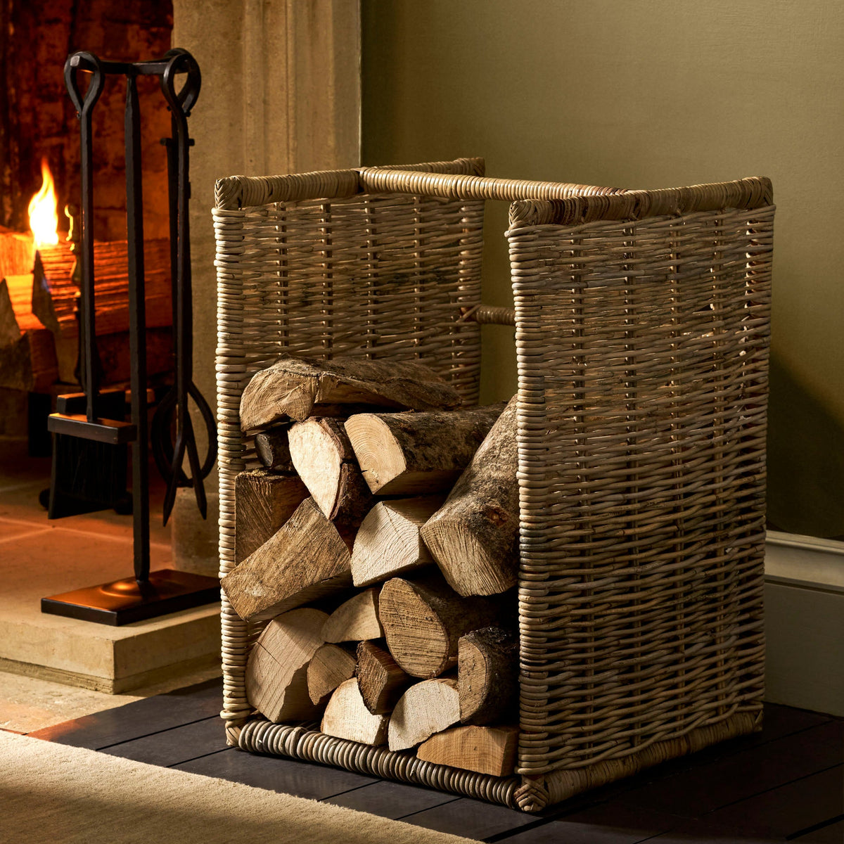 A natural woven wicker firewood storage basket filled with logs, placed on a dark hearth mat in front of a fireplace with a visible fire and metal poker.