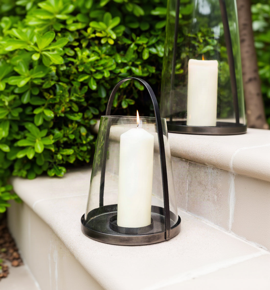 A black metal lantern with a clear glass body and a lit white pillar candle, placed on a stone ledge with green foliage in the background.