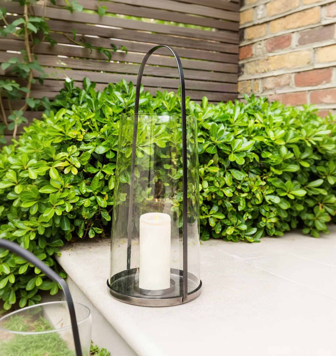 A clear glass cylindrical lantern with a black metal handle and base, holding a white pillar candle, set outdoors on a stone surface with green foliage and a wooden fence behind it.