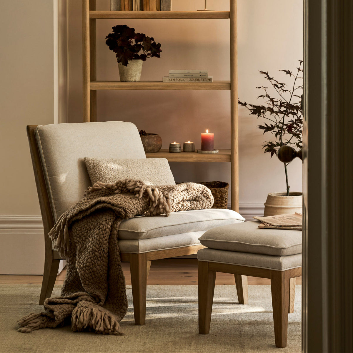 A styled interior scene showing a light wood shelving unit with books, vases, and candles, alongside a beige upholstered armchair with a throw blanket and a matching ottoman in a softly lit room.