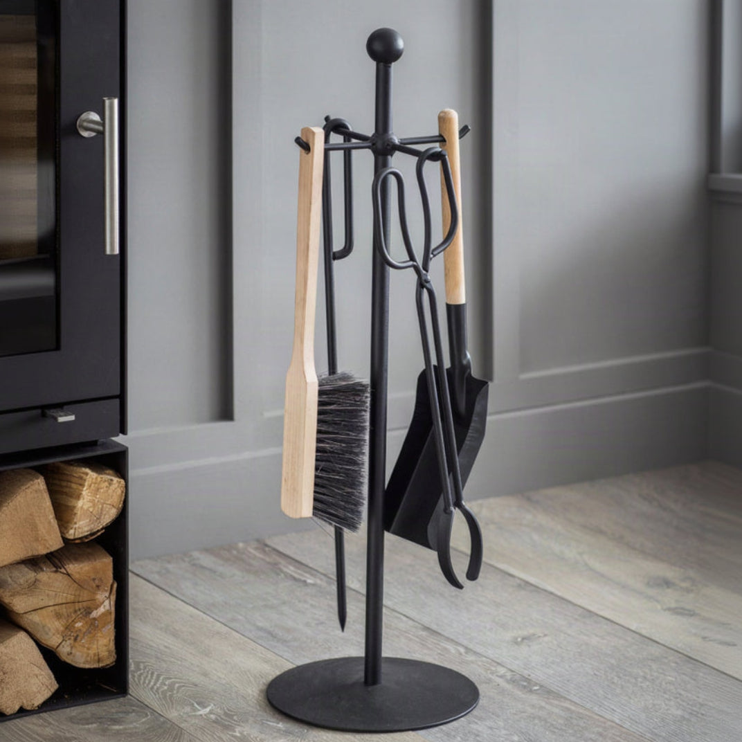 A black metal fireplace tool stand holding a brush, shovel, poker, and other fireplace tools, set on a light wood floor next to a black fireplace insert.