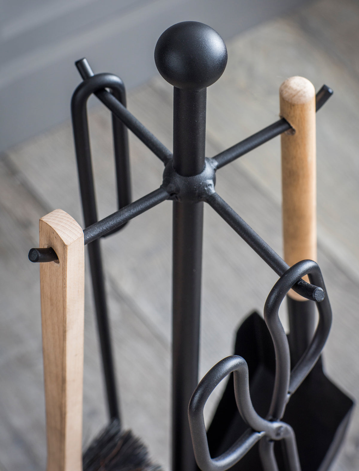 Black metal fireplace tool stand with light natural wood handles and black metal tools, photographed close-up on a light wood floor.