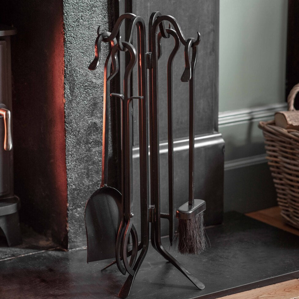 A black cast iron fireplace tool set with a matching stand, including a poker, shovel, brush, and hammer, displayed on a dark hearth in a rustic room.