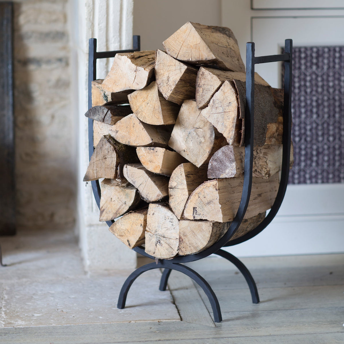 A black metal firewood rack filled with cut logs, photographed indoors near a doorway and fireplace area.