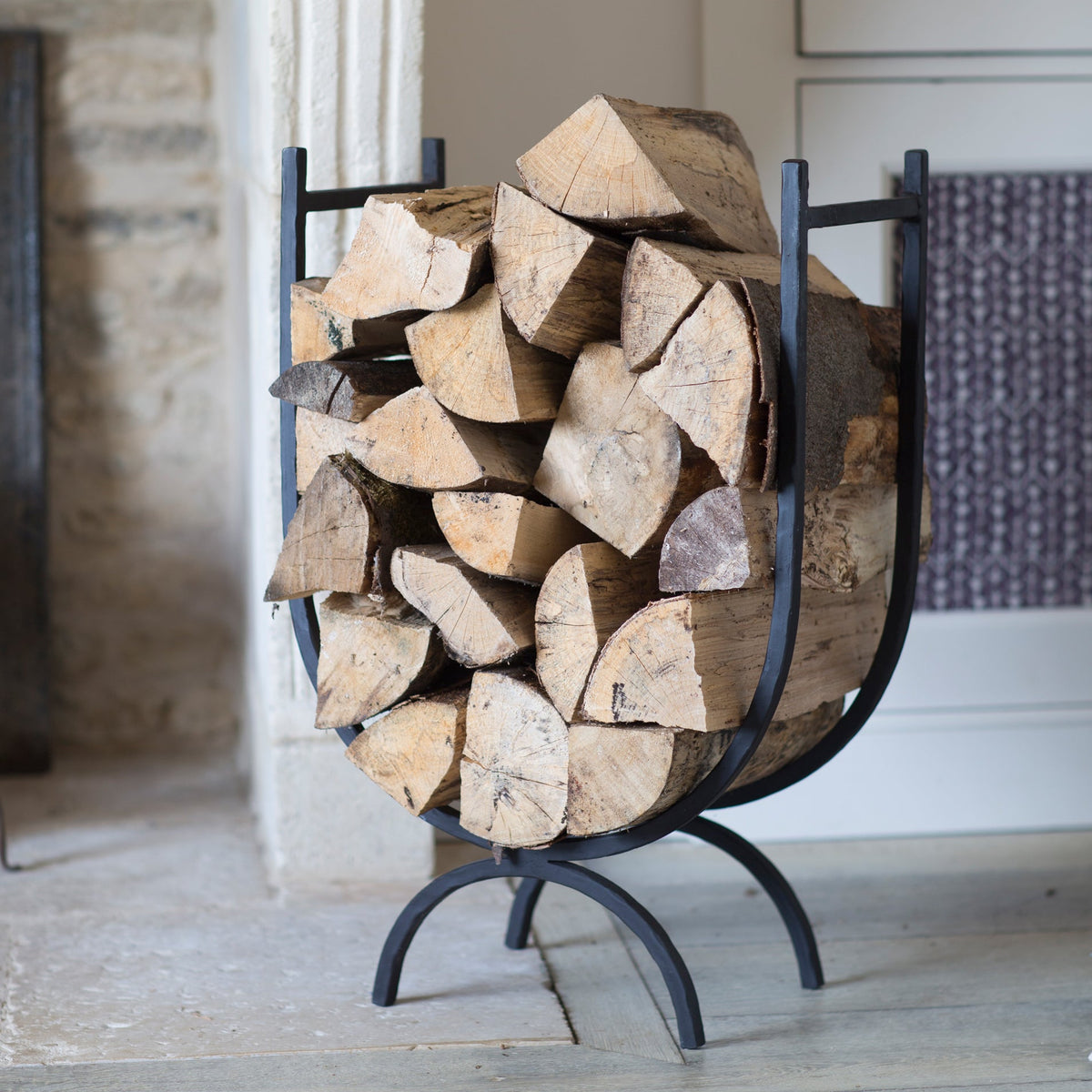 A black metal firewood rack filled with cut logs, photographed indoors near a doorway and fireplace area.