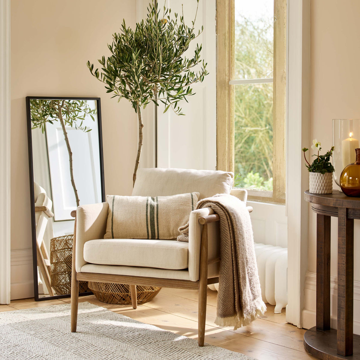 A cream upholstered armchair with a light wood frame sits in a bright, warm-toned room beside a tall window. A beige throw blanket is draped over the arm, a striped pillow rests on the seat, and a tall potted olive tree stands behind the chair.