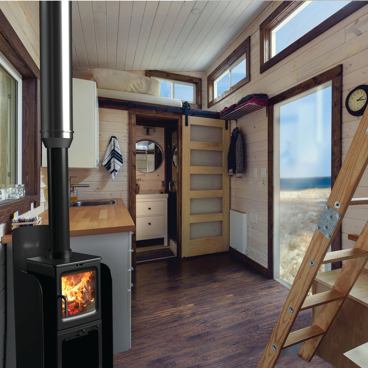 A cozy interior room with light wood-paneled walls, a black wood-burning stove with visible flames, a tall wooden ladder, and large windows looking out to a beach scene.