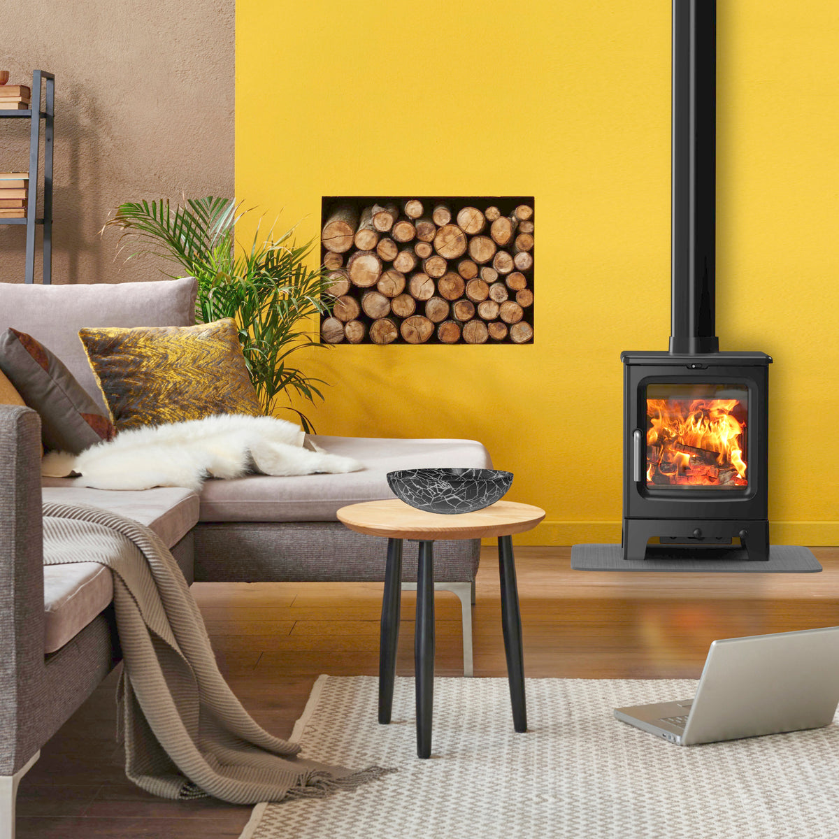 A black wood-burning stove with a visible fire, set against a bright yellow wall in a modern living room. A matching framed picture of stacked firewood hangs on the wall above the stove.