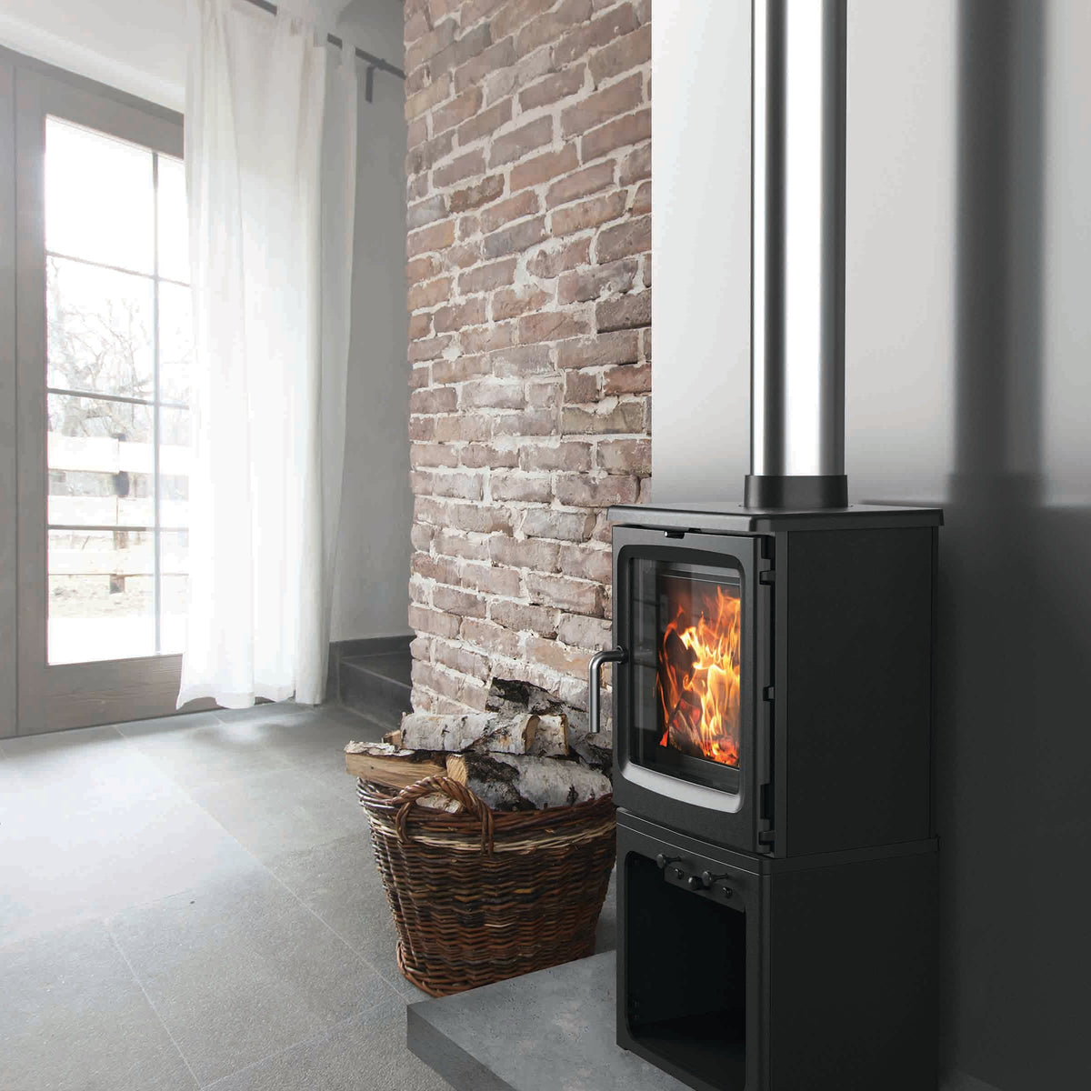 A black wood-burning stove with a visible fire, set against a brick wall in a modern room with gray tiled floors and a window with white curtains.