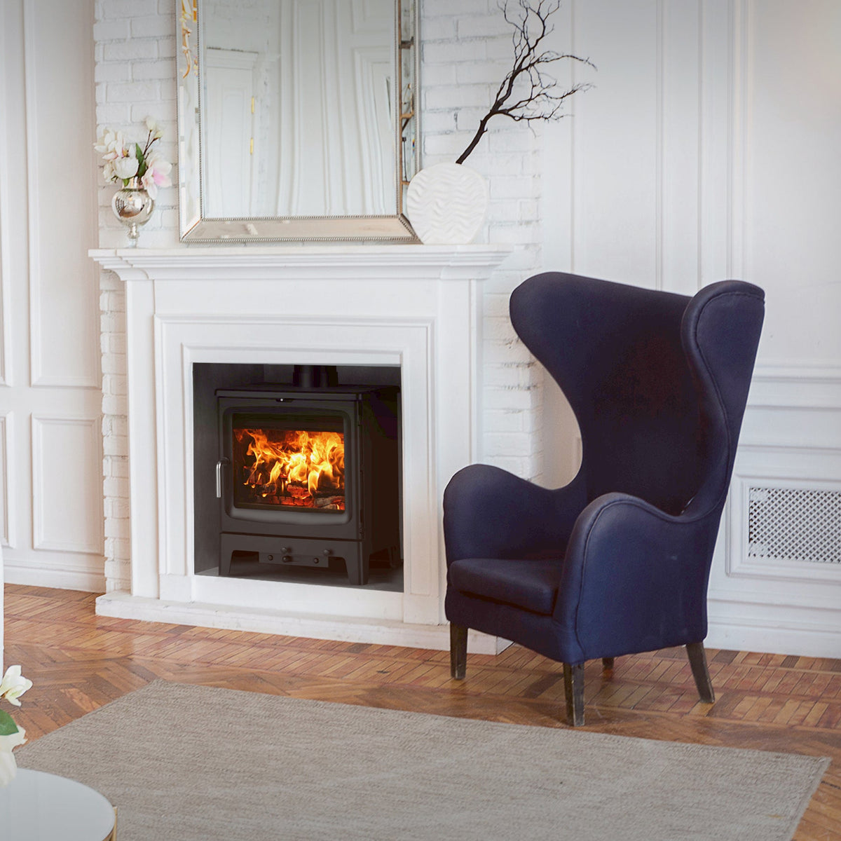A styled living room featuring a white tufted sofa, a dark blue wingback armchair, a white fireplace with a lit fire, and a tall wooden bookshelf against a white paneled wall.