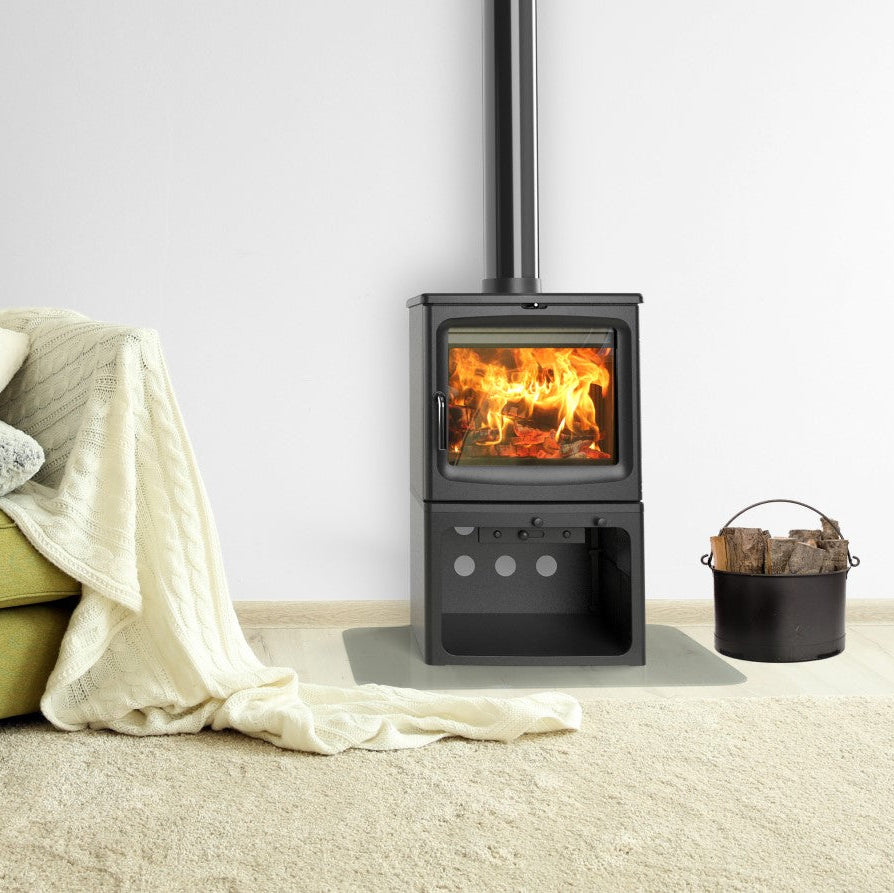 A black wood-burning stove with a visible fire, set in a cozy room next to an olive green armchair with pillows and a cream throw blanket, on a light beige rug.