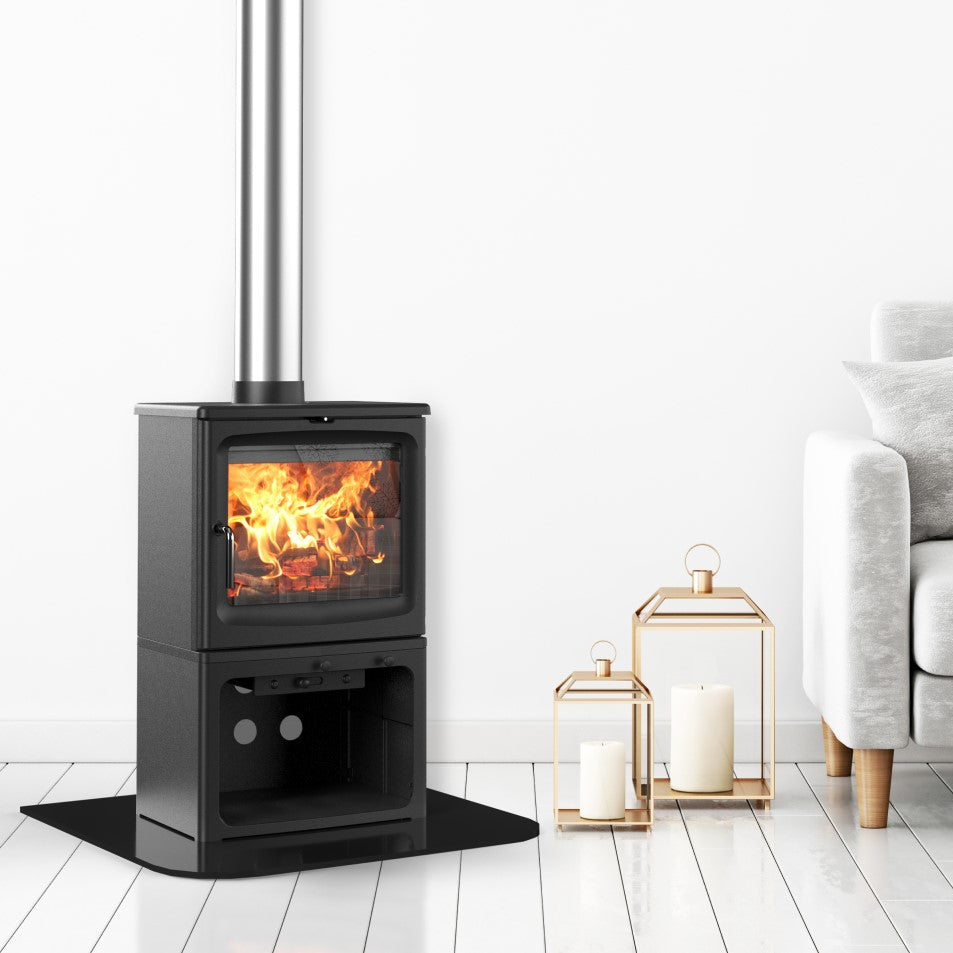 A black wood-burning stove with a visible fire and a tall silver chimney pipe, set in a modern living room next to a light gray sofa with a plaid throw blanket and decorative lanterns.