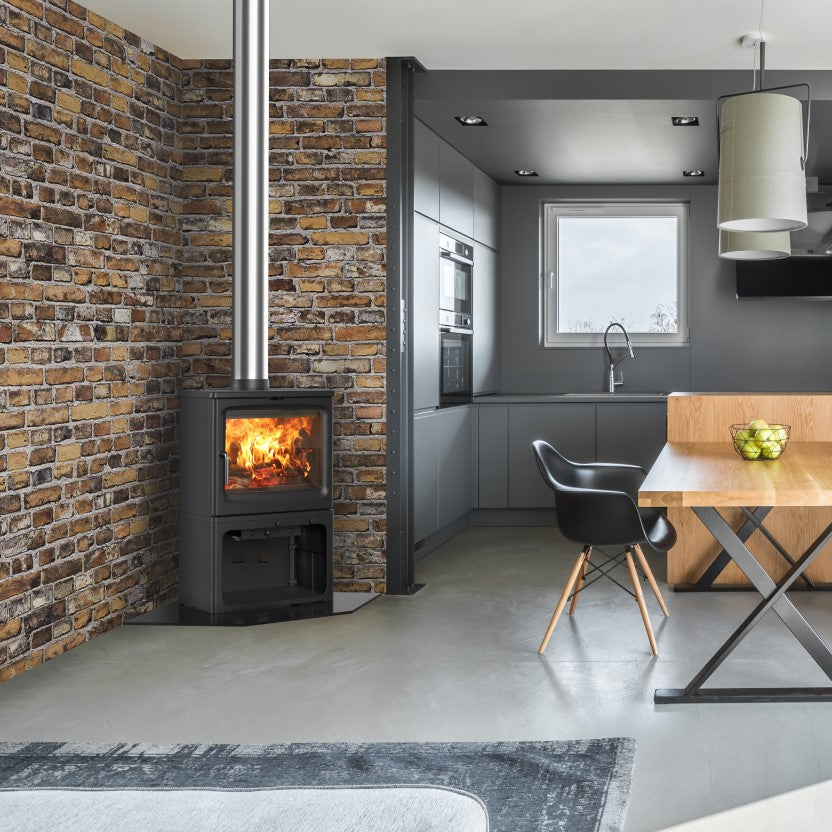 A modern interior room featuring a black wood-burning stove with a visible fire against a brick wall, a wooden dining table with black chairs, a dark gray kitchen in the background, and large glass doors leading to a balcony.