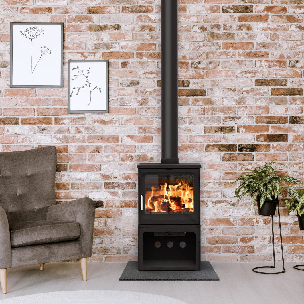 A black wood-burning stove with a visible fire, set against a brick wall in a styled living room with a gray armchair, framed wall art, and potted plants.