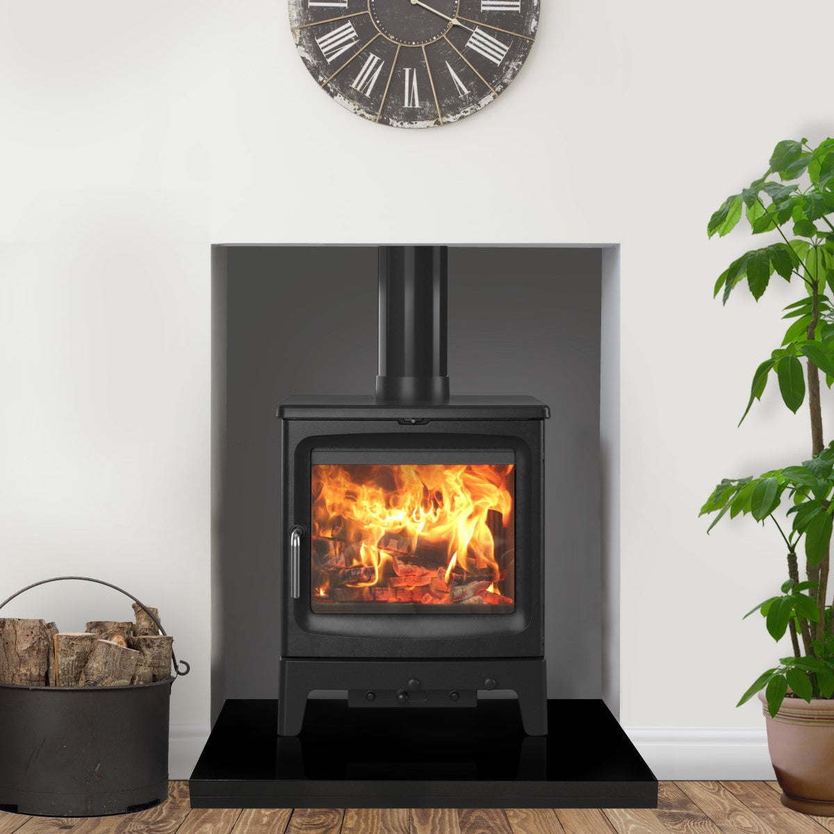 A black wood-burning stove with a glowing fire inside, set against a dark gray fireplace surround in a staged room. A round wall clock hangs above, with a potted plant on the right and a bucket of logs on the left.