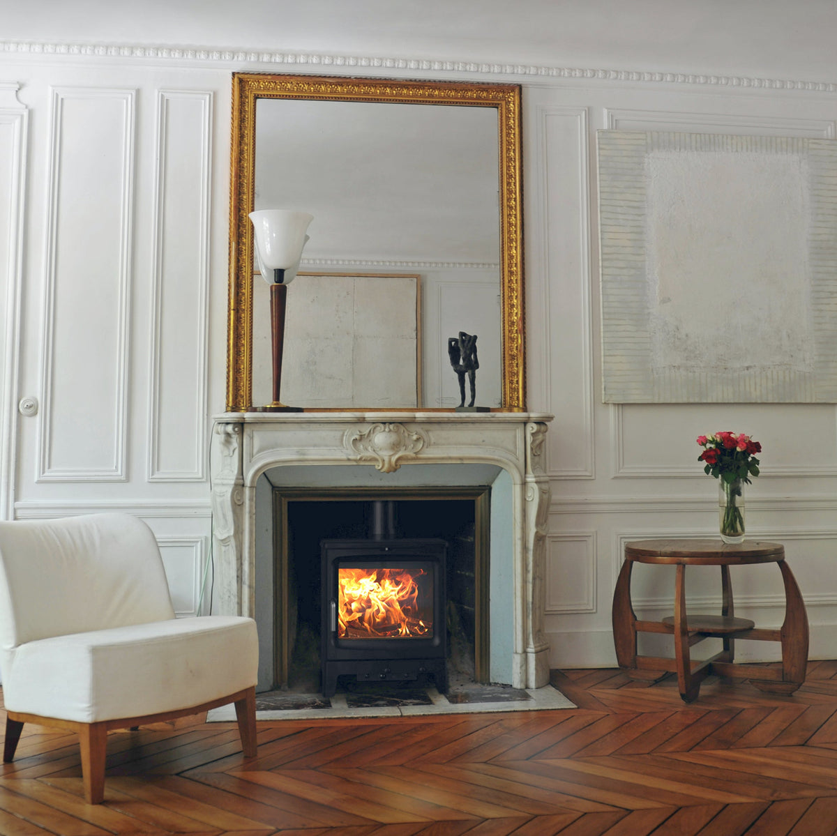 A classic French interior room with white paneled walls, a central fireplace with a gold mirror above, a white armchair on the left, and a small wooden side table with red flowers on the right, set on a herringbone wood floor.