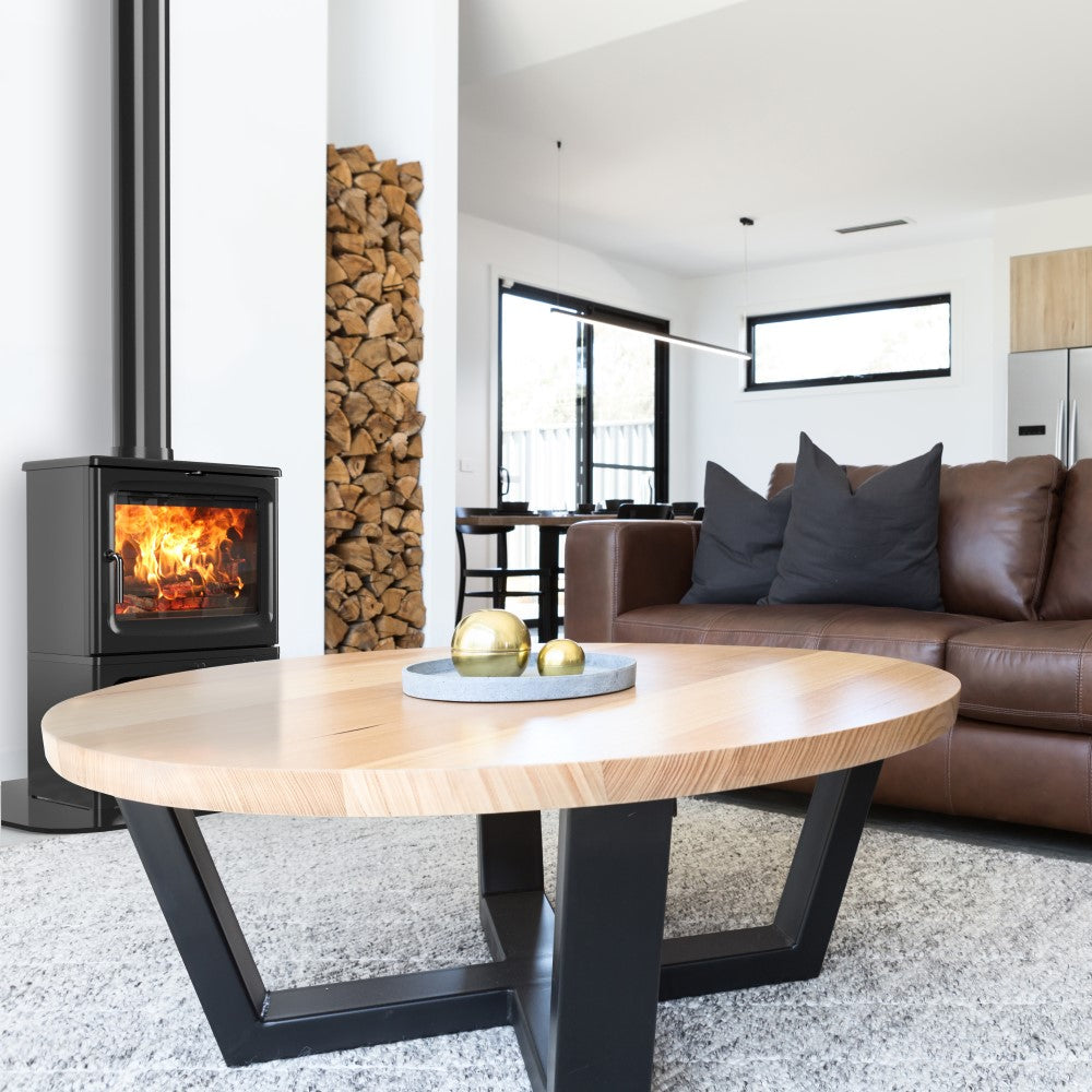 A modern living room featuring a brown leather sofa with dark gray pillows and a black-framed round coffee table. To the left is a black wood-burning stove with a visible fire, set next to a stacked wood column.