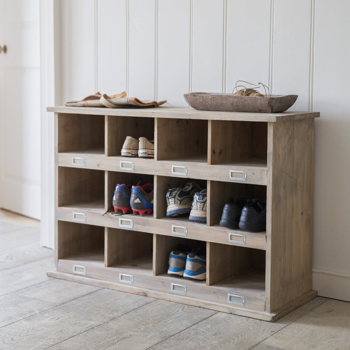 A light natural wood shoe storage cabinet with multiple cubbies, shown in a bright room with white paneled walls and light wood flooring. Several pairs of shoes are placed inside the cubbies, and a woven tray sits on top.