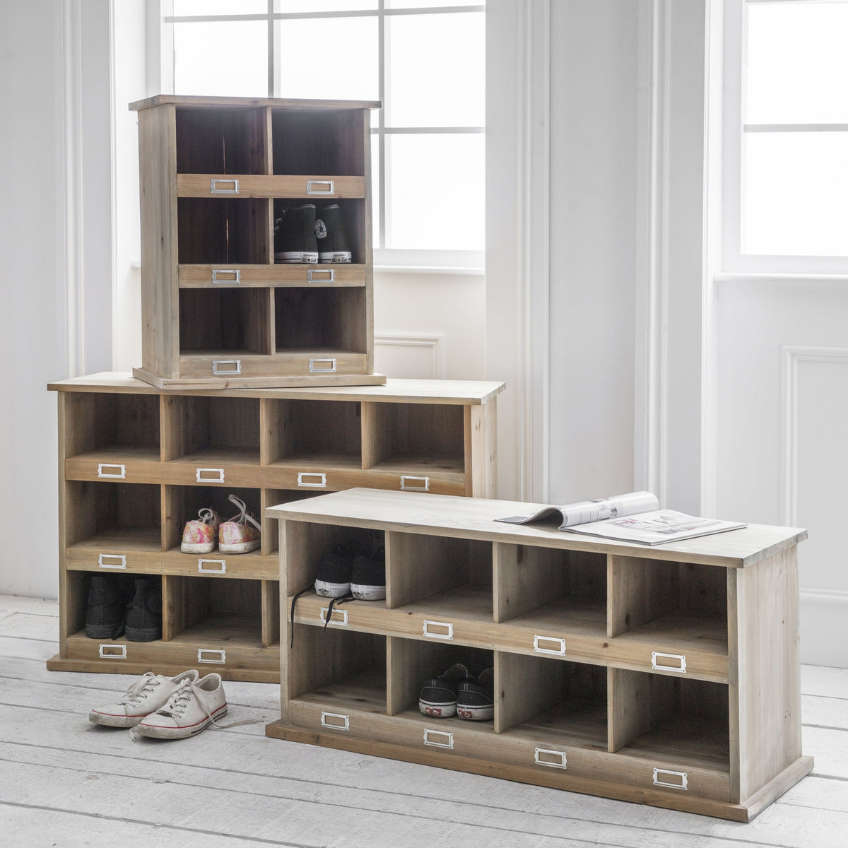 A set of light natural wood shoe lockers and a matching bench arranged in a bright white room with large windows. The lockers include cubby shelves and small labeled drawers, with several pairs of shoes stored inside.